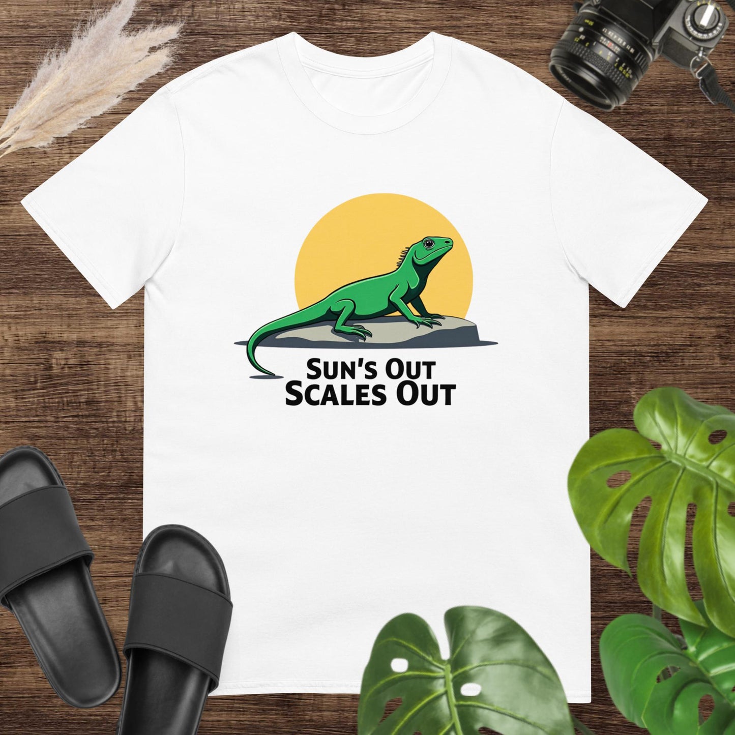 White t-shirt featuring a green lizard on a rock with a sun and the text “Sun’s Out Scales Out.”