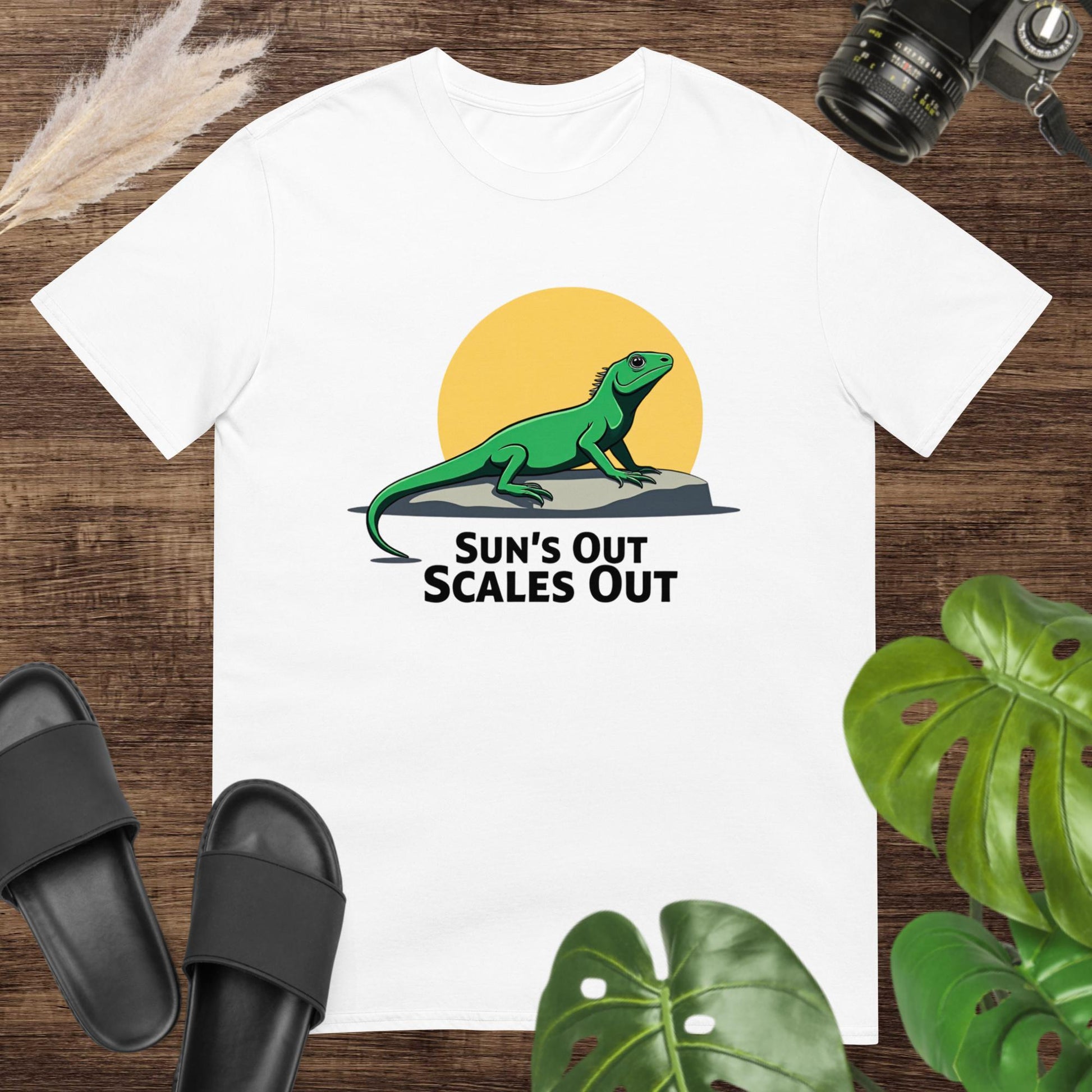 White t-shirt featuring a green lizard on a rock with a sun and the text “Sun’s Out Scales Out.”